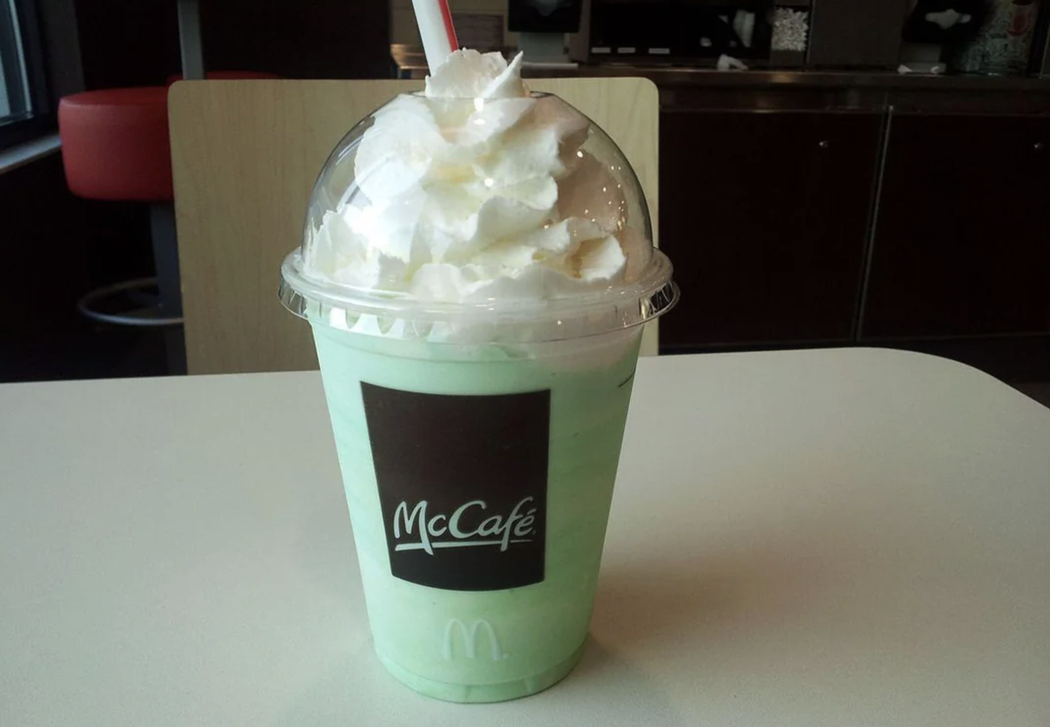 A McCafé Shamrock Shake in a clear plastic cup with whipped cream on top, sitting on a white table in a McDonald's restaurant. The shake is light green in color and has a red-striped straw.