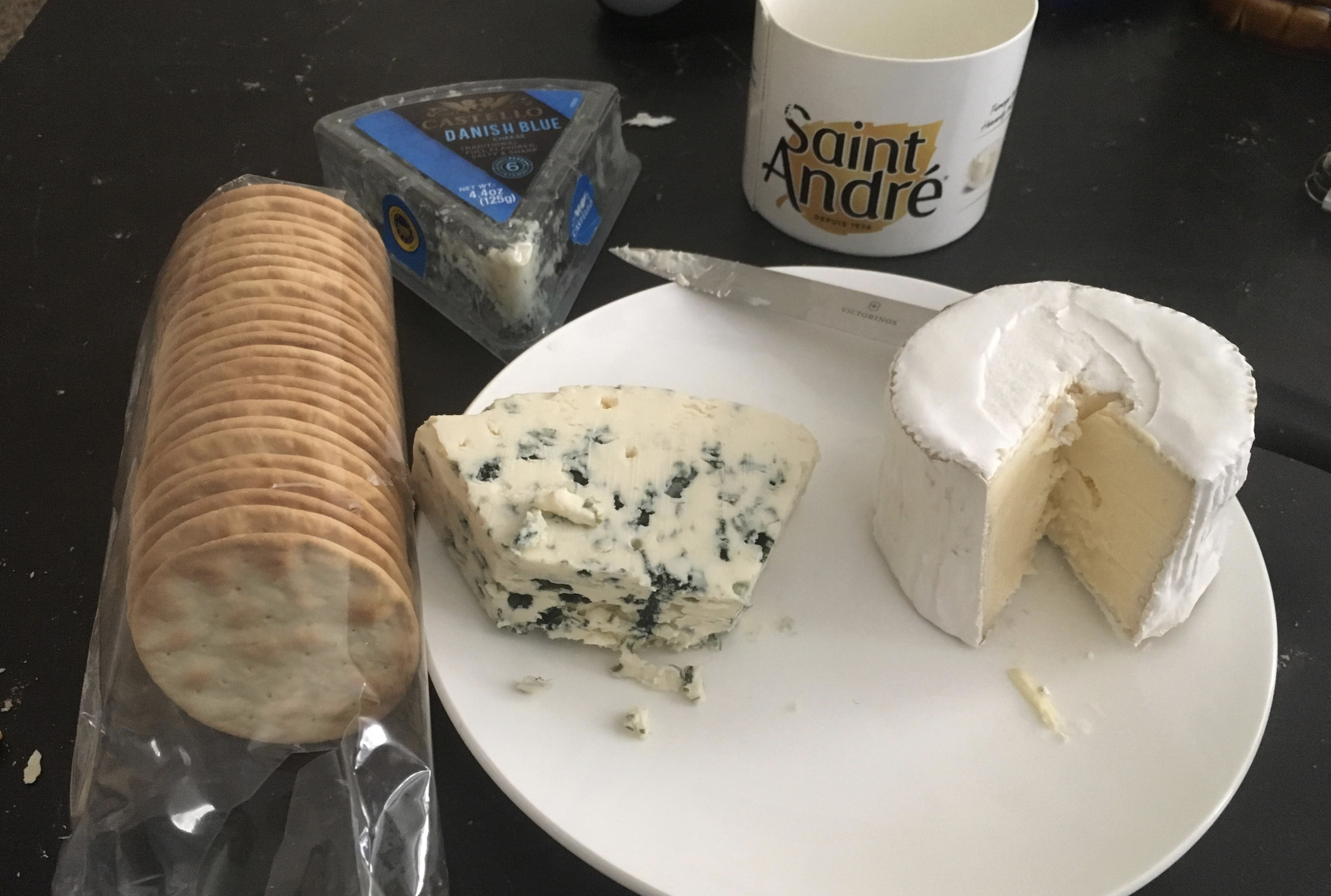 A plate with round crackers, a wedge of blue cheese, and a wheel of soft cheese. Nearby are a knife, a mug labeled "Saint André," and a package of Danish blue cheese on a dark surface.