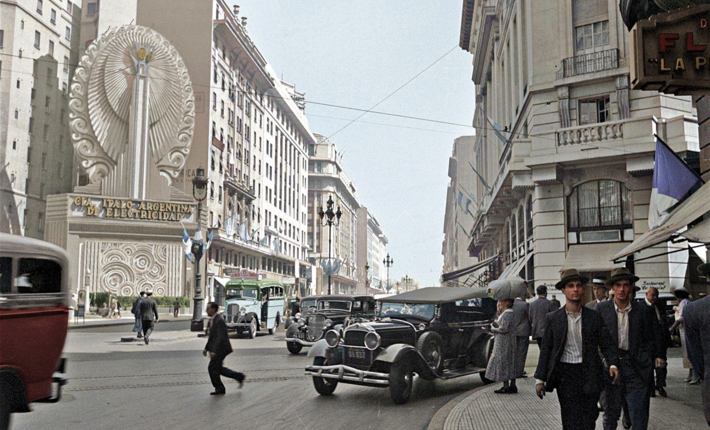 A busy city street in the early 20th century features vintage cars, people in suits and hats, and buildings decorated with Argentine flags. A large, ornate façade advertises electricity at the intersection.