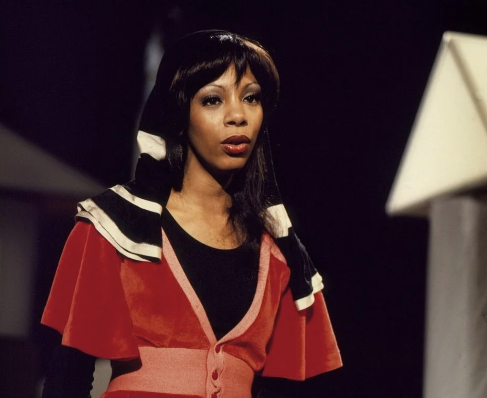 A woman with long dark hair wears a red and black outfit with a matching hood. She stands indoors against a dark background with a white geometric shape on the right side.
