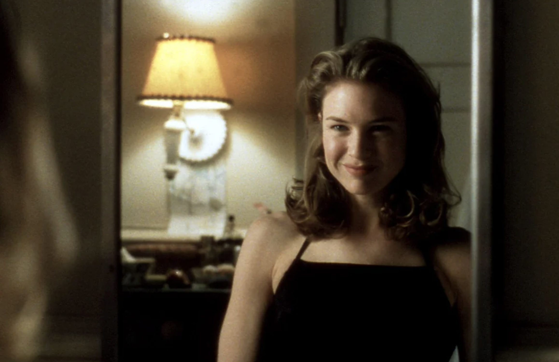 A woman with wavy brown hair in a black dress smiles while looking into a mirror, with a softly lit lamp and blurred decor in the background.