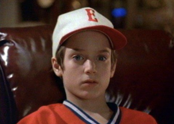 A young boy wearing a white baseball cap with a red brim and a red letter "E," along with a red shirt, sits on a brown leather chair, looking ahead with a neutral expression.