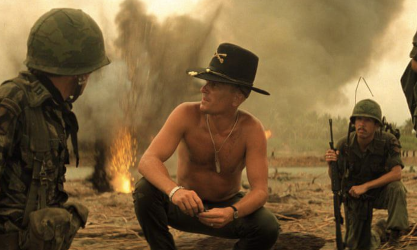 Three soldiers in military gear kneel and crouch on a smoky battlefield; one is shirtless and wears a black cavalry hat and sunglasses. An explosion and heavy smoke rise in the background.