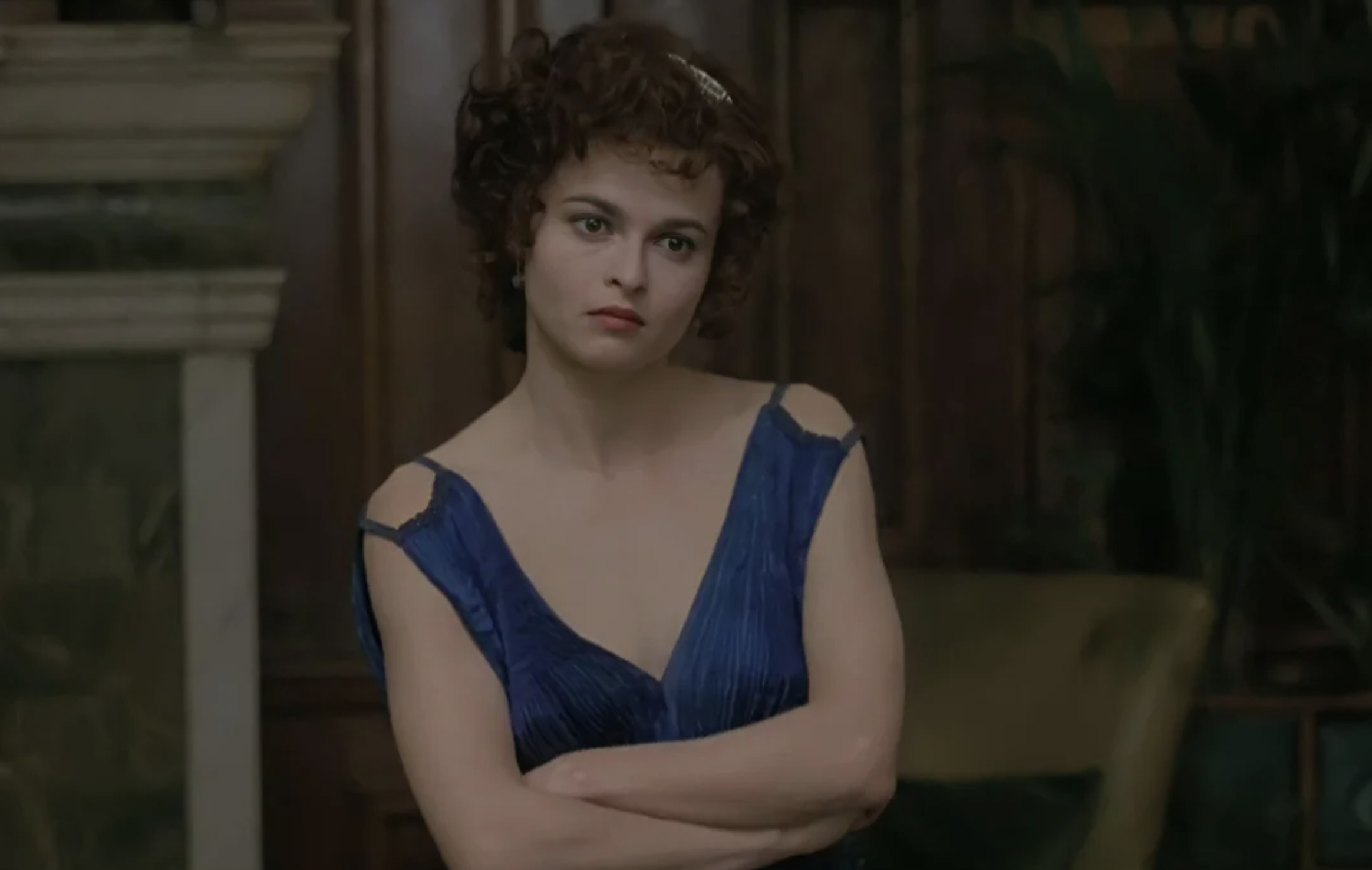 A woman with short, curly brown hair stands with her arms crossed, wearing a blue sleeveless dress. She has a neutral expression and is in a room with dark wood paneling and green furniture.