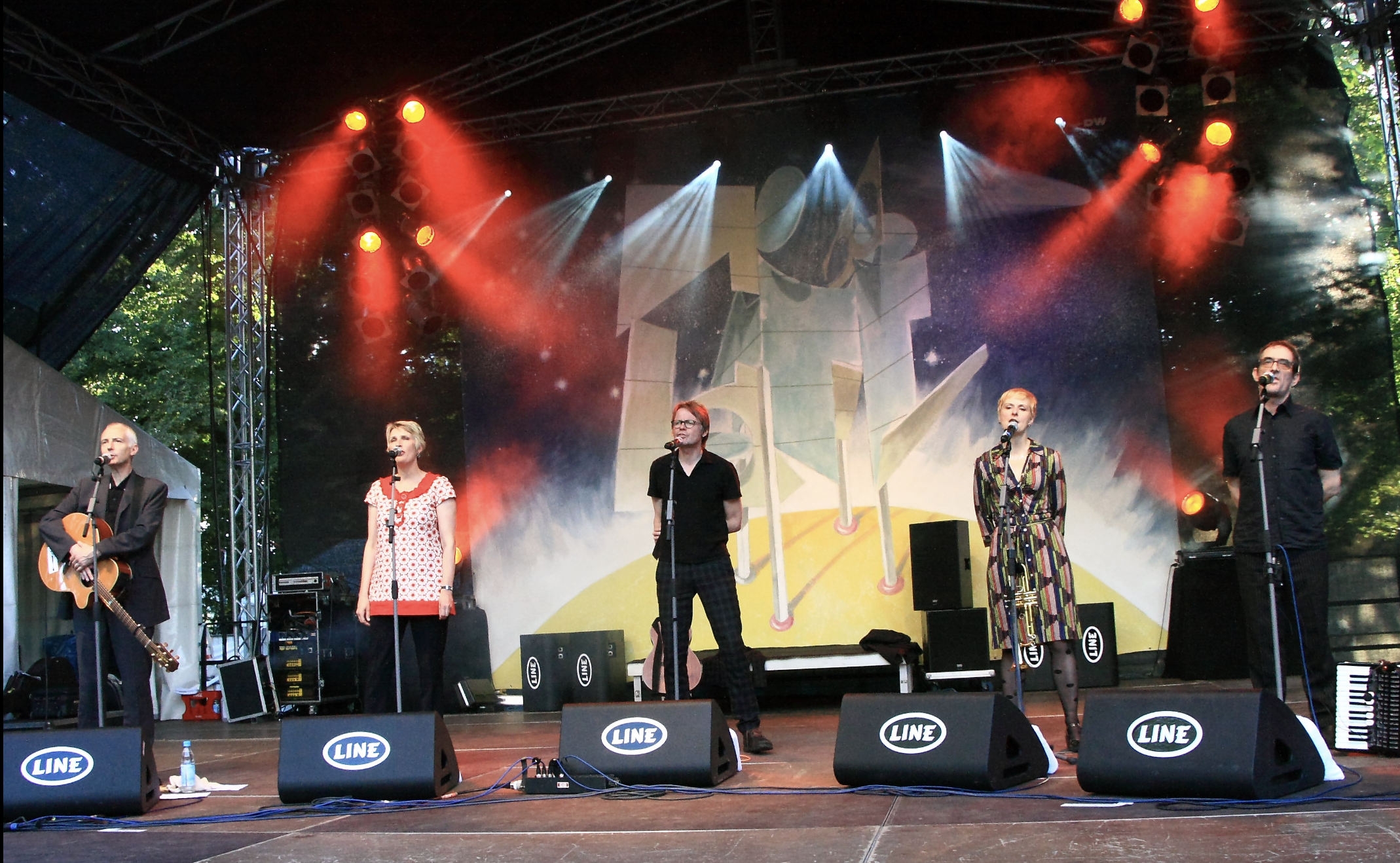 Five musicians stand on an outdoor stage with microphones, colorful lights, and a painted backdrop behind them. One plays guitar, and the others sing. Various equipment and speakers are visible in the scene.