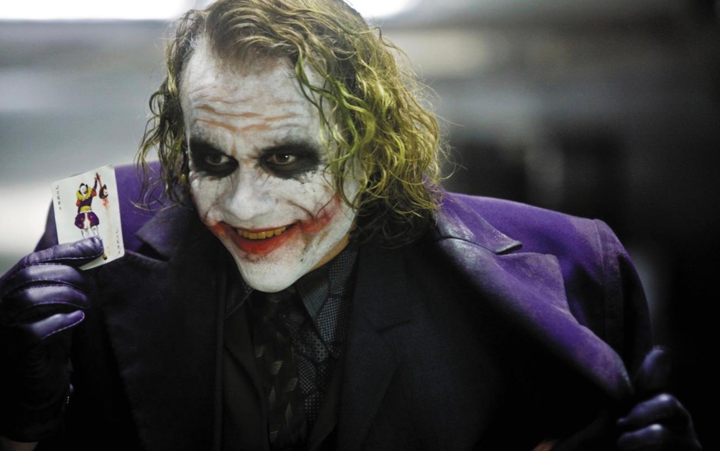 A person in white face paint with dark eye makeup, red smeared lipstick, green-tinted hair, and a purple coat holds up a Joker playing card, smiling menacingly. The background is dim and blurry.