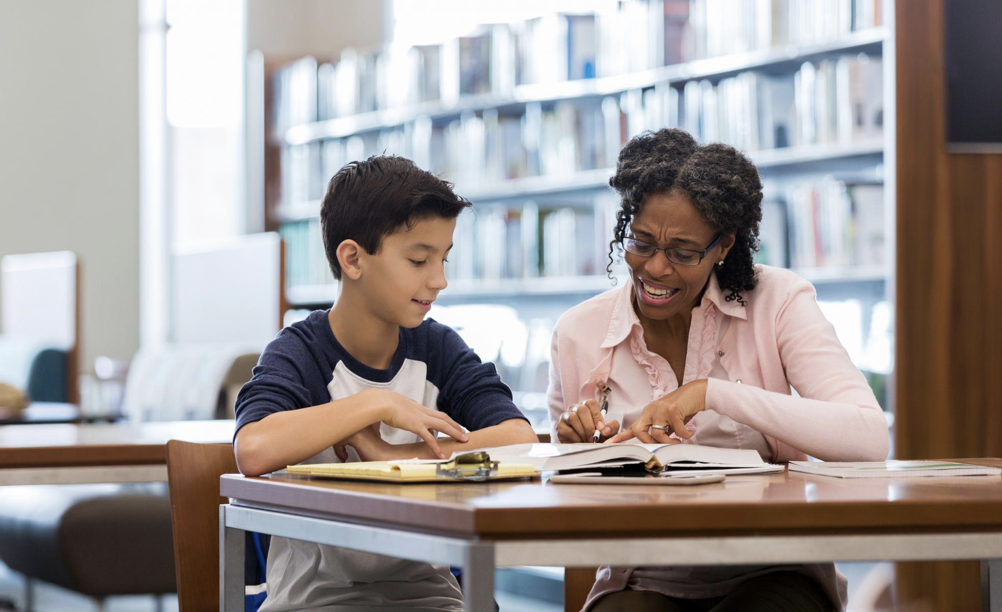 A woman and a boy sit at a library table, reading and discussing a book together. The woman smiles while pointing at the book, and the boy listens attentively. Shelves of books are visible in the background.