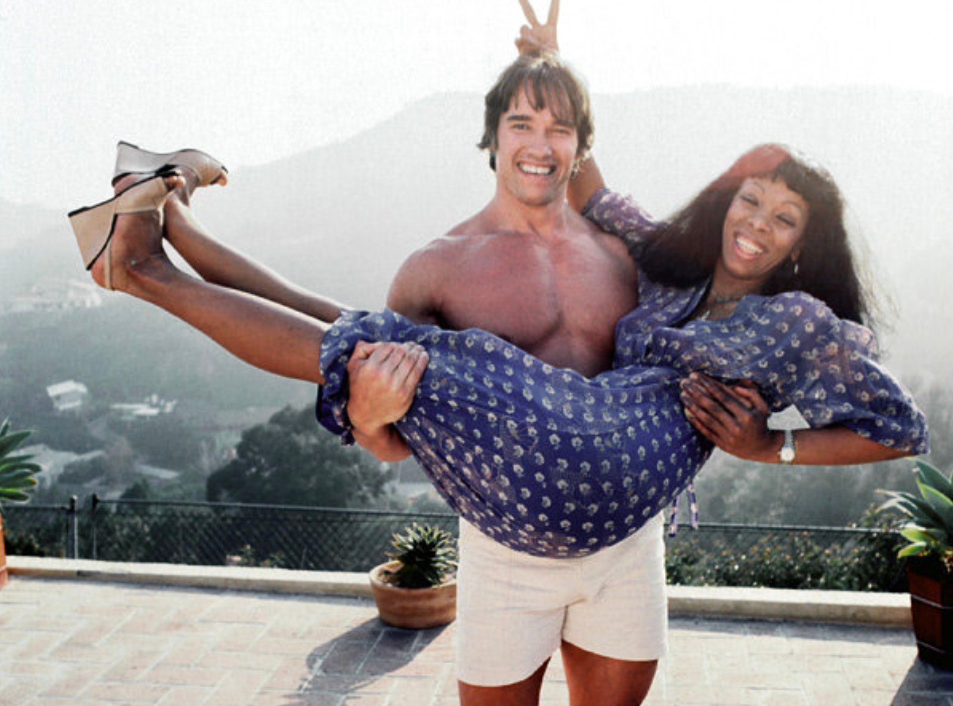 A smiling shirtless man in white shorts playfully lifts a laughing woman in a blue dress, with mountains and potted plants in the background outdoors. The man gives the woman bunny ears with his fingers.