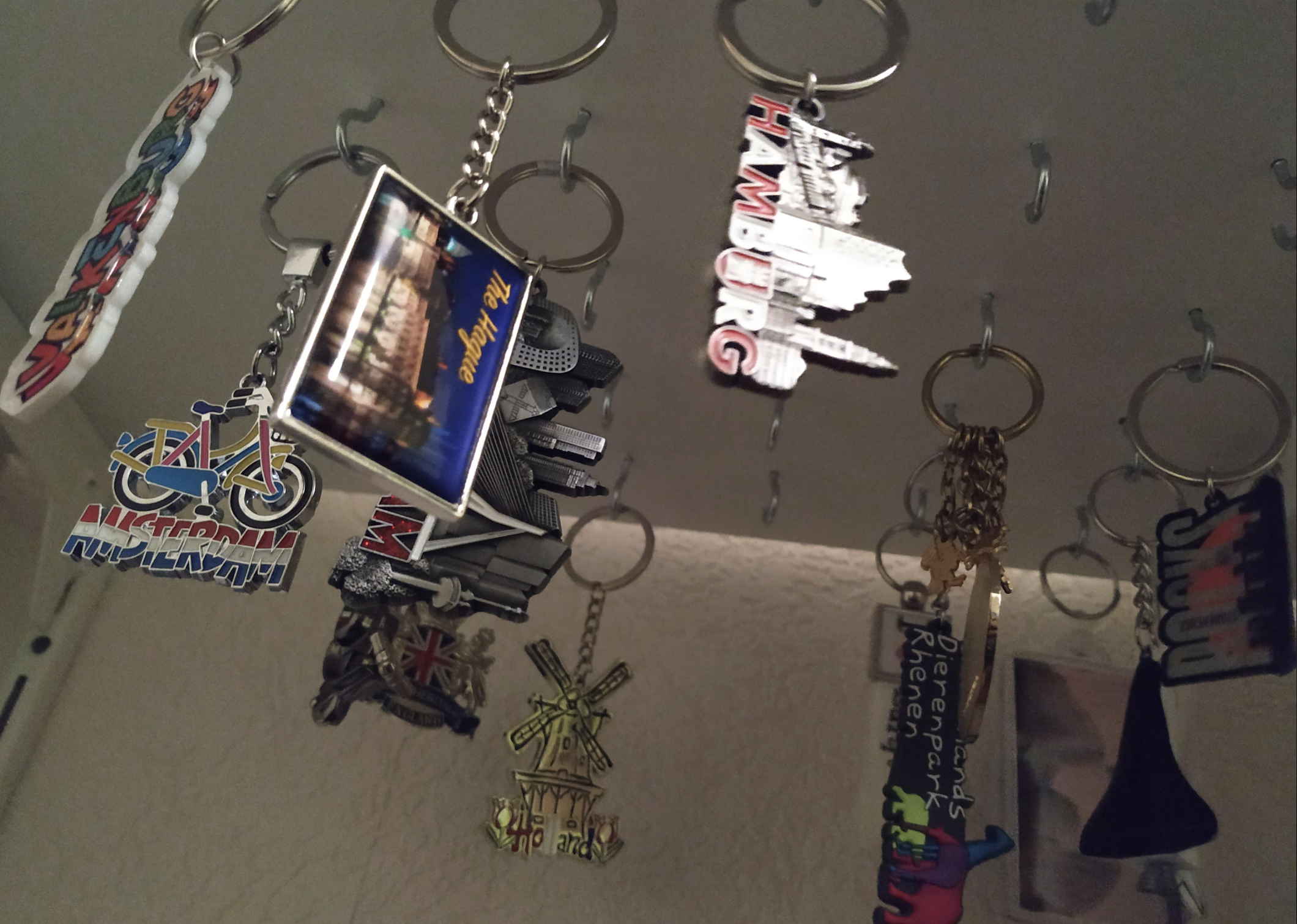 A collection of colorful souvenir keychains from various cities, including a windmill, a motorcycle, and landmarks, hang from hooks under a shelf against a beige wall.