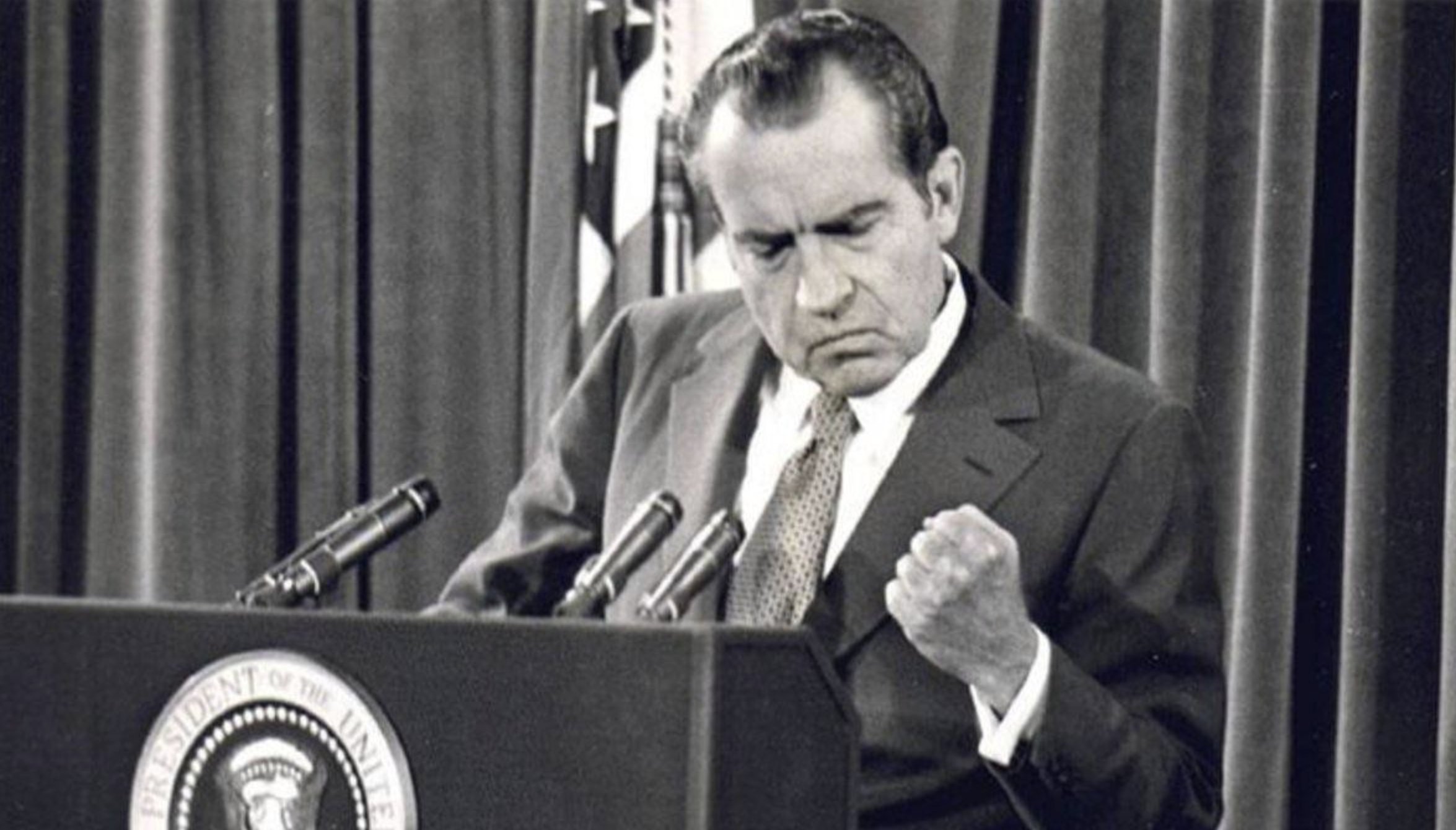 A man in a suit stands at a podium with microphones, making a fist with his right hand and a determined expression. An American flag and a presidential seal are visible in the background.