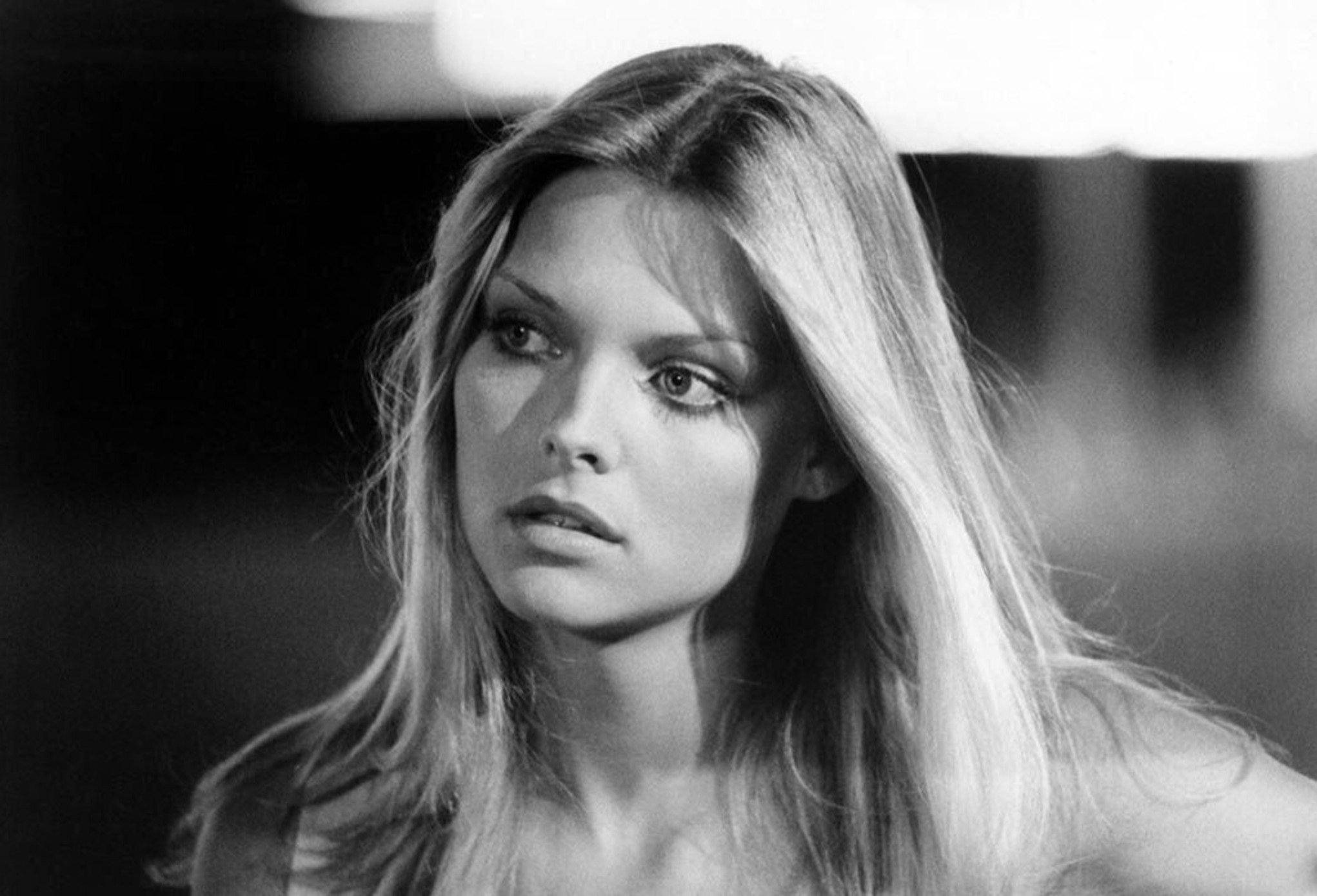 Black and white photo of a young woman with long, straight blonde hair, looking slightly to her left with a neutral expression. She wears a light top and the background is softly blurred.