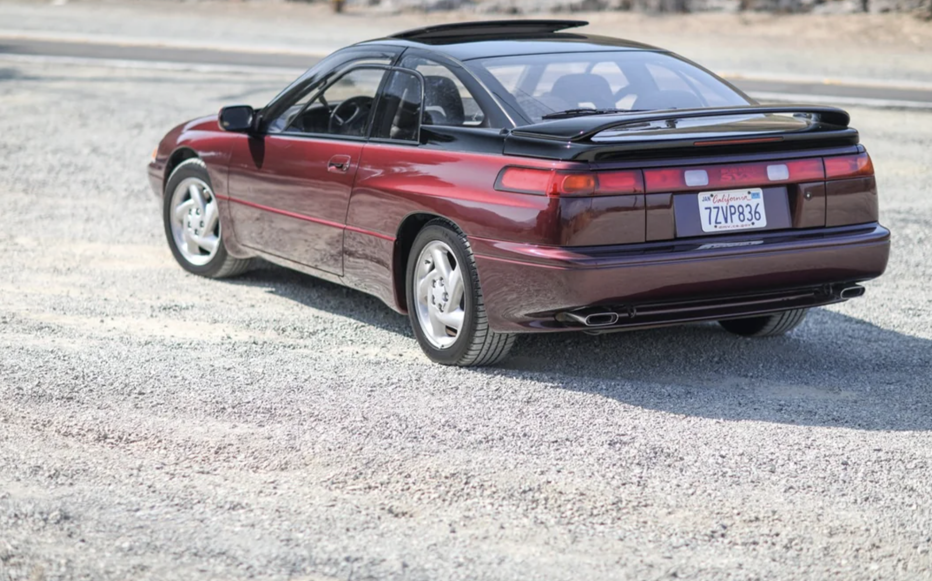 A maroon Subaru SVX coupe with a sunroof and spoiler is parked on a gravel surface, viewed from the rear left side. The car has a California license plate.