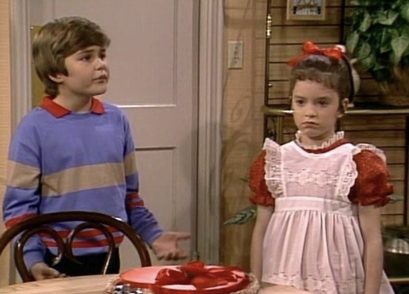 A young boy in a blue and red striped sweater talks to a young girl in a red dress with a white apron. The girl looks serious; both stand indoors near a table with a red cake on it.