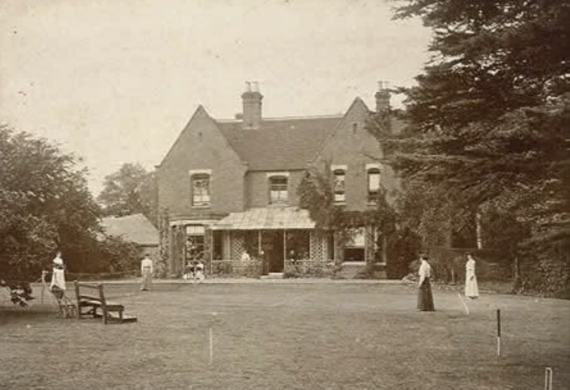 A sepia-toned photograph shows people playing lawn games in front of a large brick house covered in ivy, surrounded by trees and a garden. The scene appears to be from the late 19th or early 20th century.