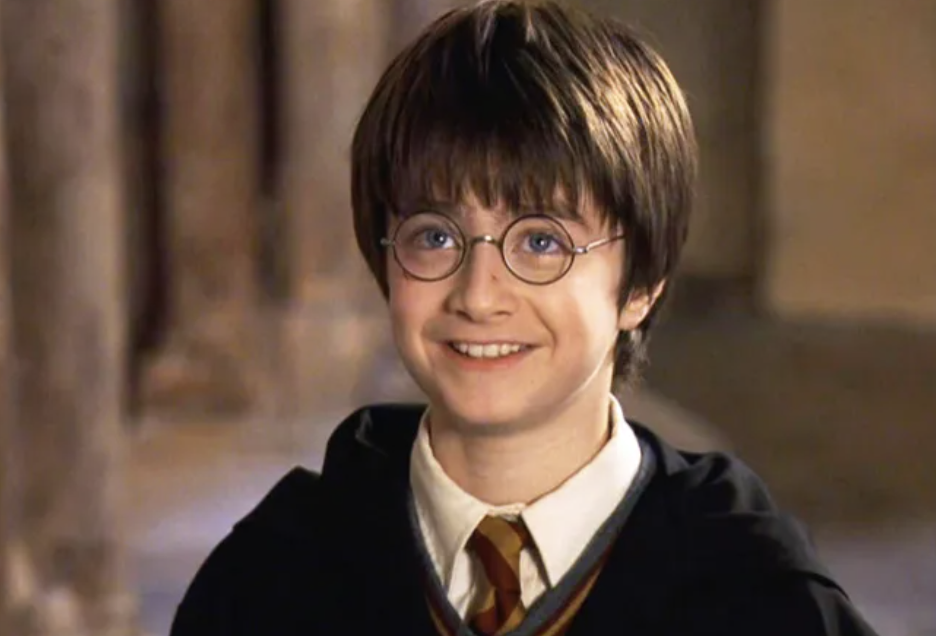 A young boy with round glasses, short brown hair, and a lightning-shaped scar on his forehead smiles while wearing a black cloak and a red-and-gold striped tie in a stone-walled setting.