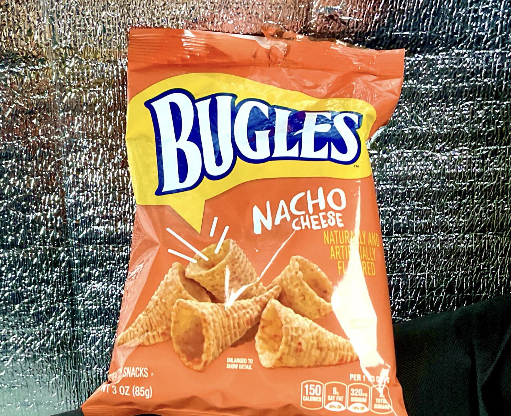 A bag of Bugles Nacho Cheese flavored snacks against a silver textured background. The package features images of cone-shaped Bugles chips and bold blue and white "Bugles" branding.