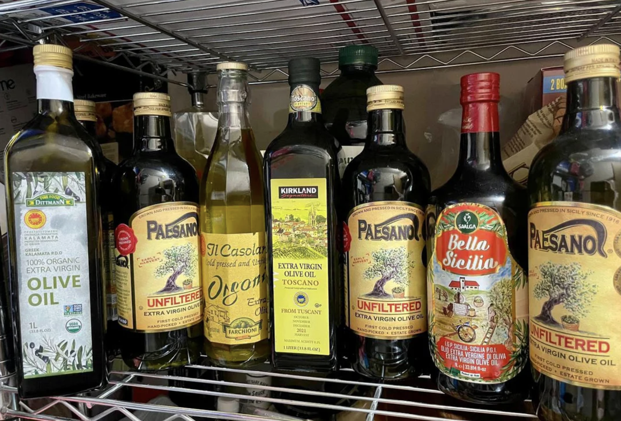 Several bottles of olive oil, including brands like Paesano, Kirkland, Bella Sicilia, Osporos, De Cecco, and O-Live, are arranged on a wire pantry shelf. The labels are colorful and feature various designs.