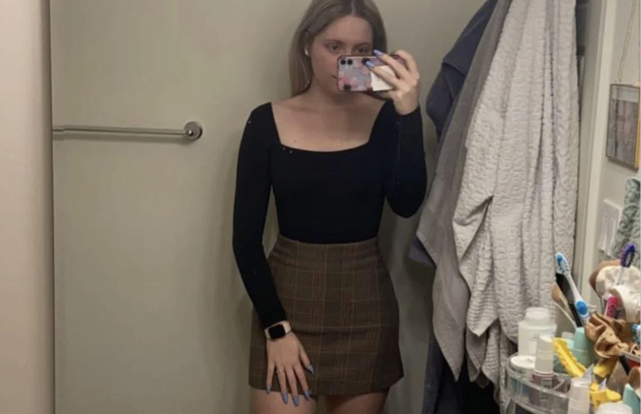 A person stands in a bathroom taking a mirror selfie. They wear a black long-sleeve top and a brown plaid skirt. Various toiletries and towels are visible on shelves to the right.