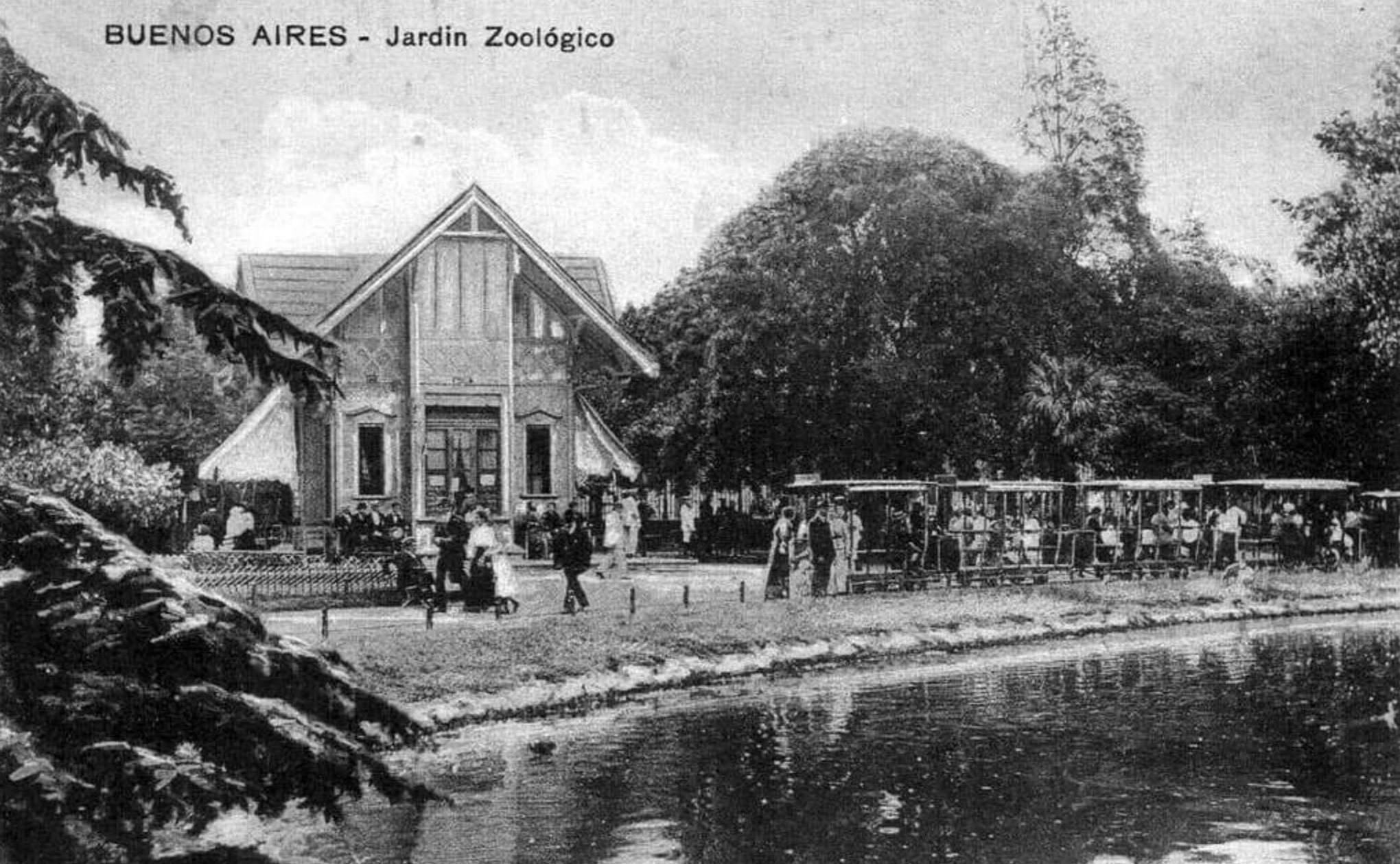Black and white photo of the Buenos Aires Zoo. People gather near a pond in front of a large building, with trees around. A small open-air train with passengers is visible on the right side.