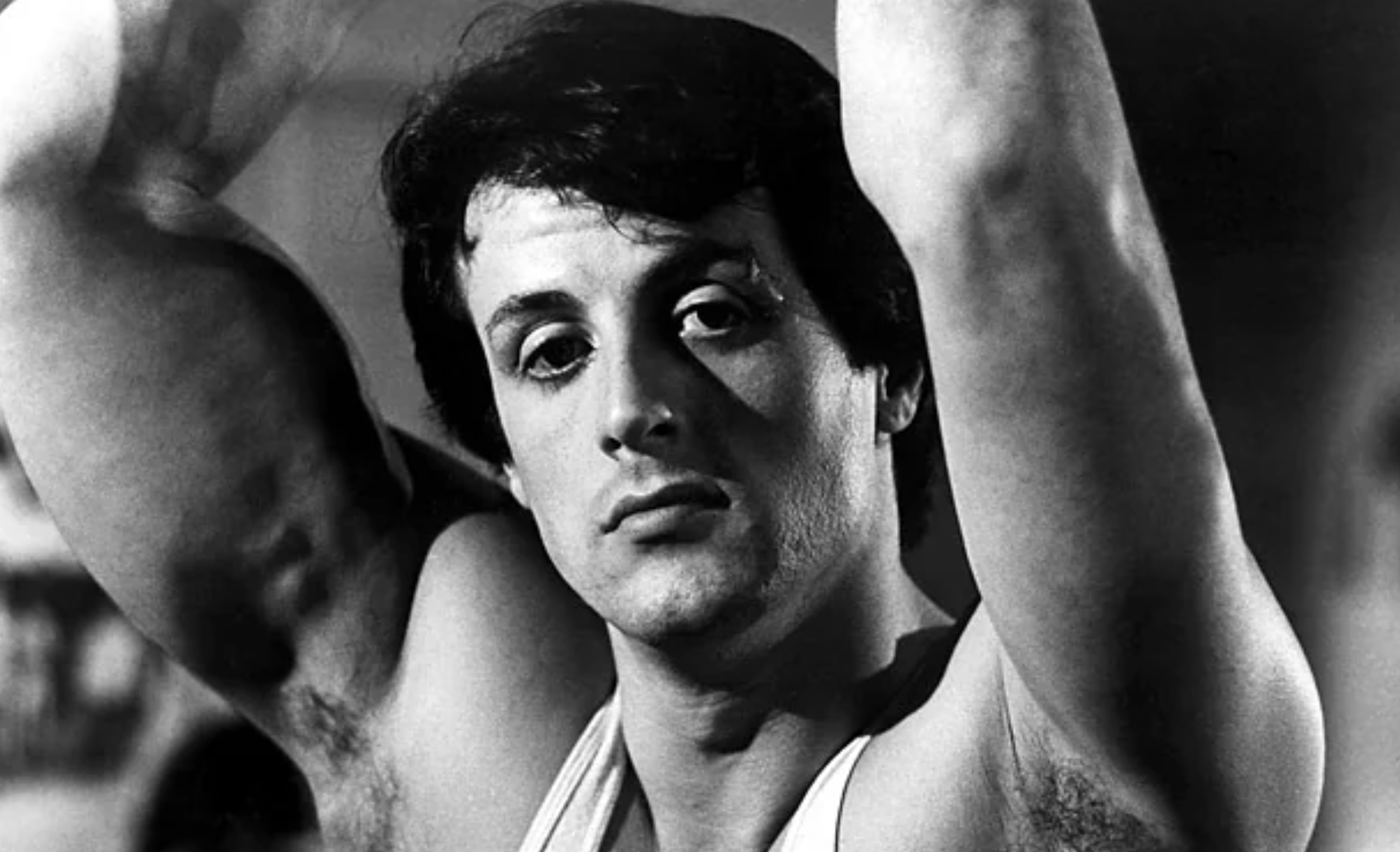 A black-and-white photo of a young man with dark hair, wearing a tank top, raising his arms above his head, and looking directly at the camera with a neutral expression.