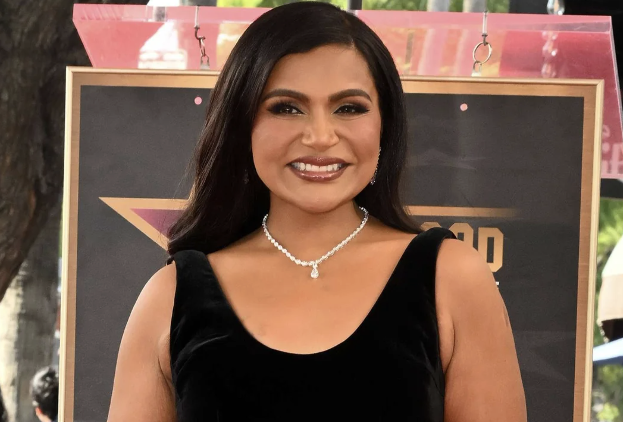 A woman with long dark hair, wearing a black sleeveless dress and a silver necklace, smiles while standing outdoors in front of a decorative backdrop.