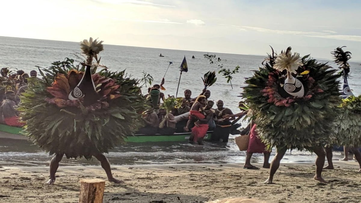 Two people in elaborate costumes made of leaves and masks dance on a beach while a group of people arrive in decorated boats in the background, all against the backdrop of the ocean at sunset.