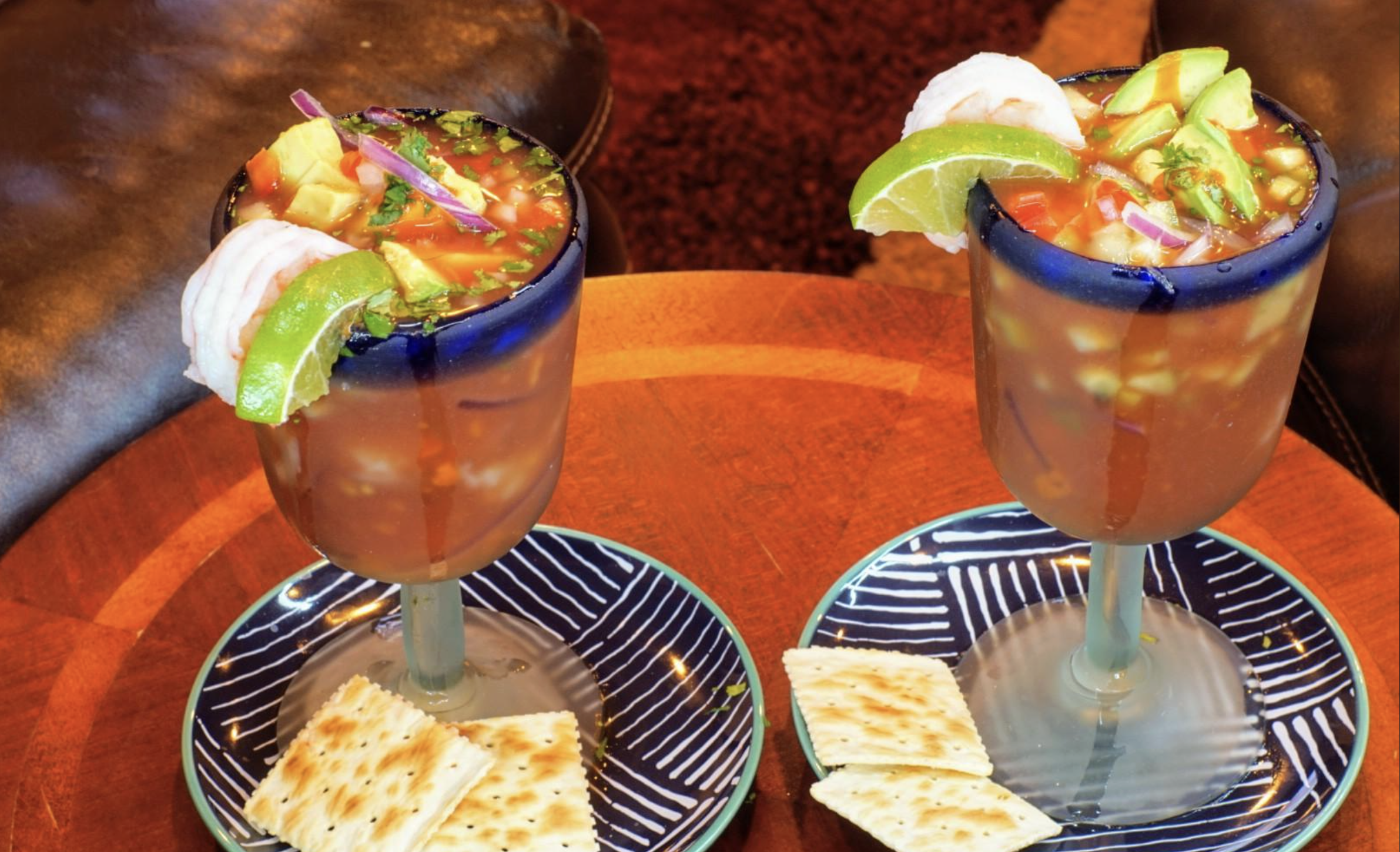 Two goblets filled with shrimp cocktail, garnished with lime, avocado, and red onion, sit on blue-striped plates with saltine crackers beside each glass on a wooden table.