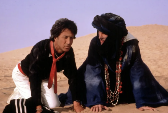 Two men kneel on sand in a desert. One wears a black jacket, white pants, and red scarf. The other wears a blue robe, large beaded necklaces, and a dark headscarf, both looking intently ahead. Sand dunes and clear sky are in the background.