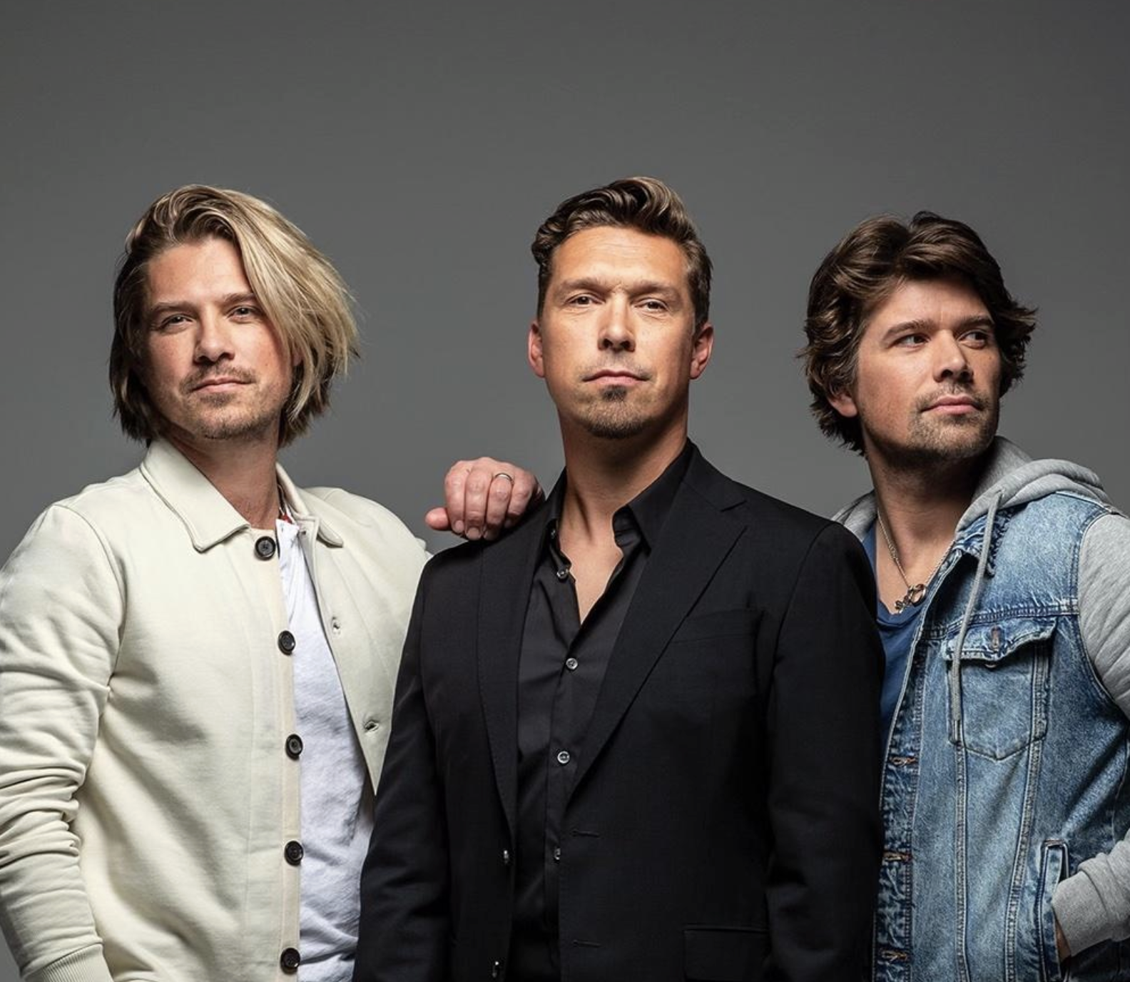 Three men with light skin stand together against a gray background. The man in the center wears a black suit, while the men on either side wear casual jackets—one in white, the other in denim. They look confidently at the camera.