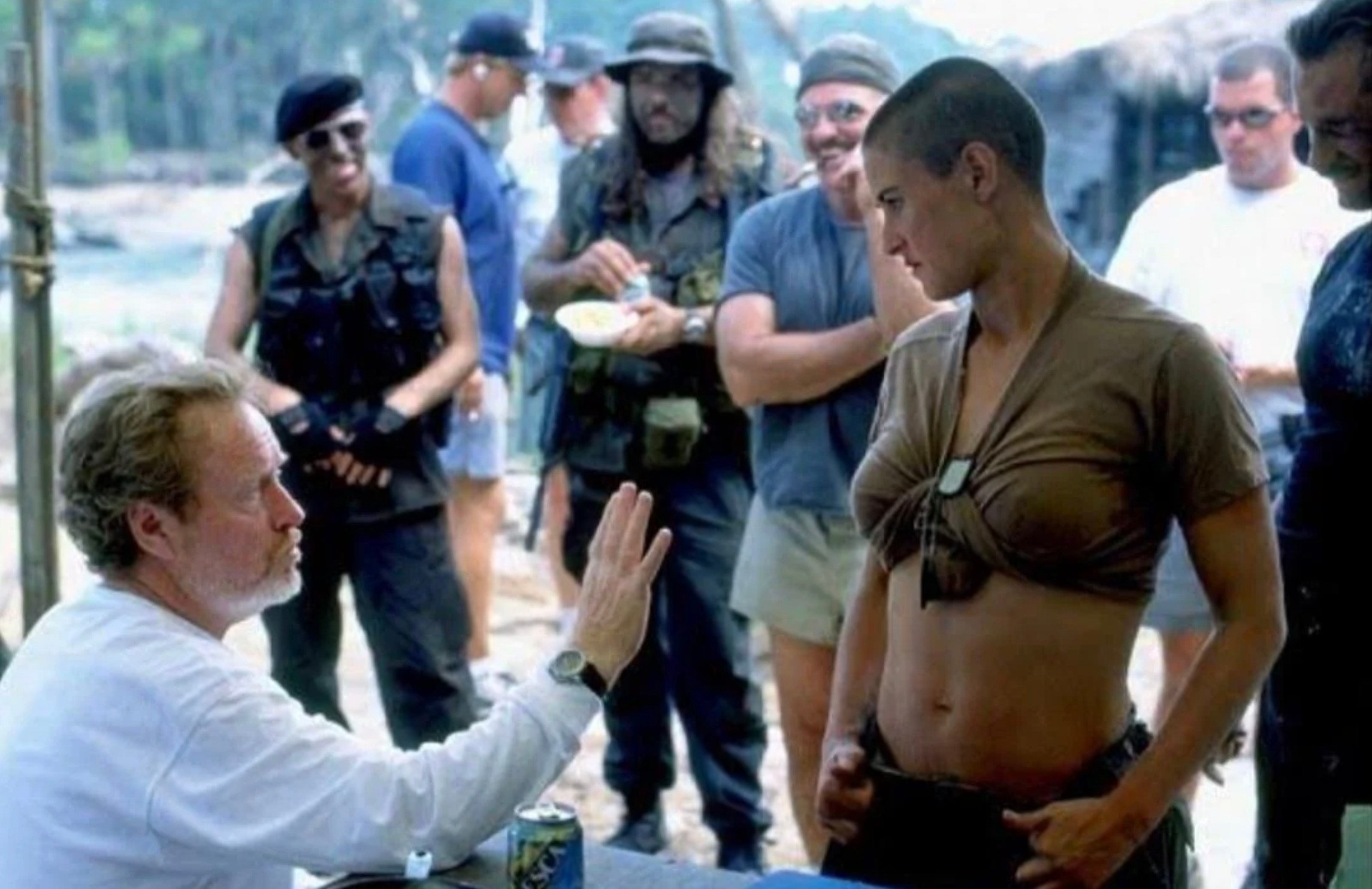 A director sits and gestures while speaking to an actor with a shaved head and a cropped shirt; crew members in military-style costumes stand and watch on a film set outdoors.
