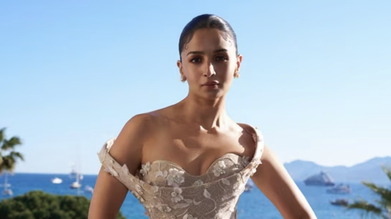 A woman in an off-shoulder, floral-patterned dress stands outdoors with her hand on her hip. The background shows a clear blue sky, palm trees, the sea, and boats in the distance.