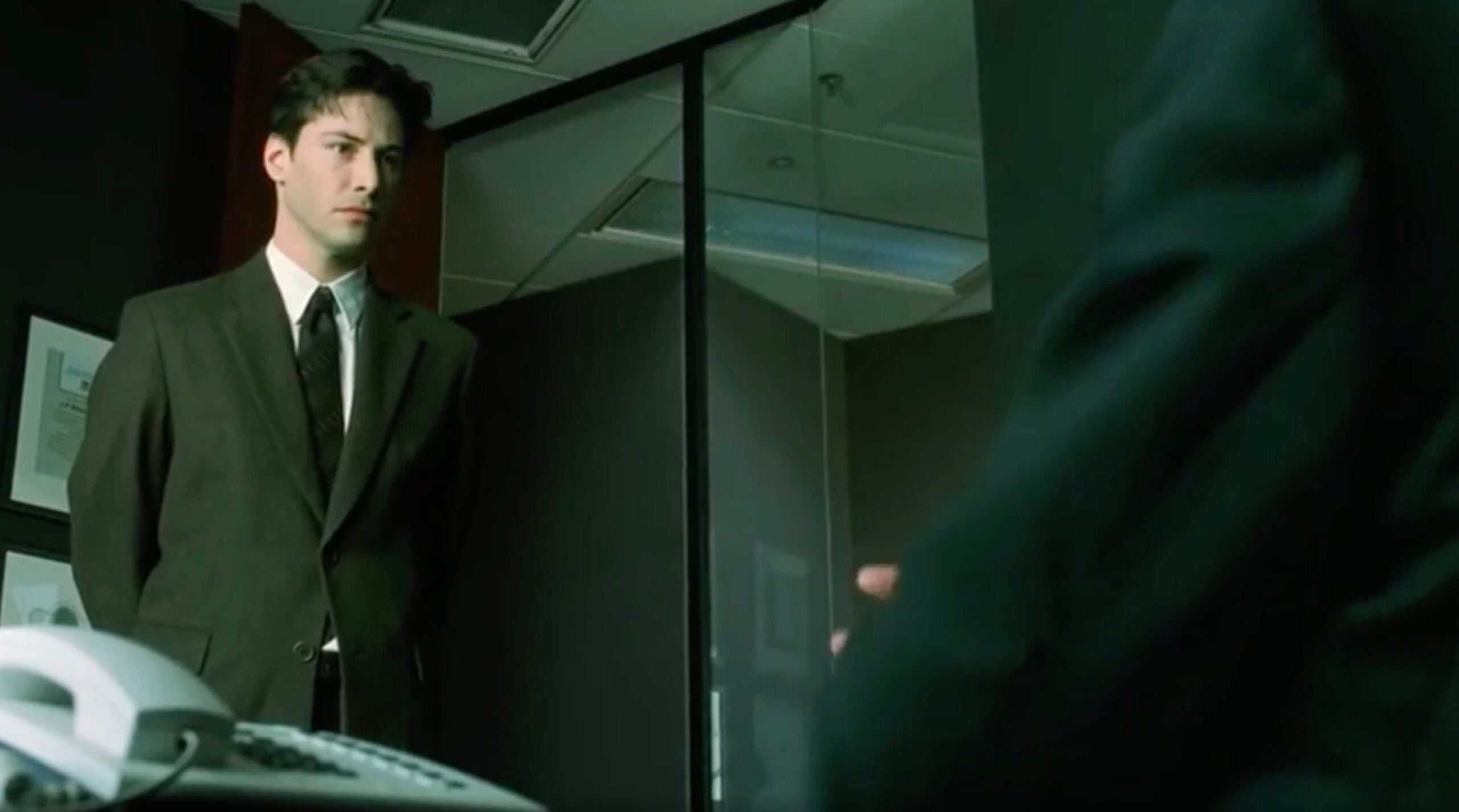 A young man in a dark suit stands nervously in an office, facing someone seated at a desk. The office has glass walls, framed pictures, and a white office phone in the foreground.