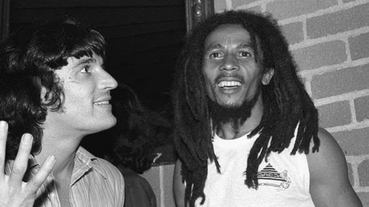 Two men in a candid black-and-white photo; one smiles at the camera wearing a sleeveless shirt with long dreadlocks, while the other faces him, gesturing with one hand. A brick wall is visible in the background.