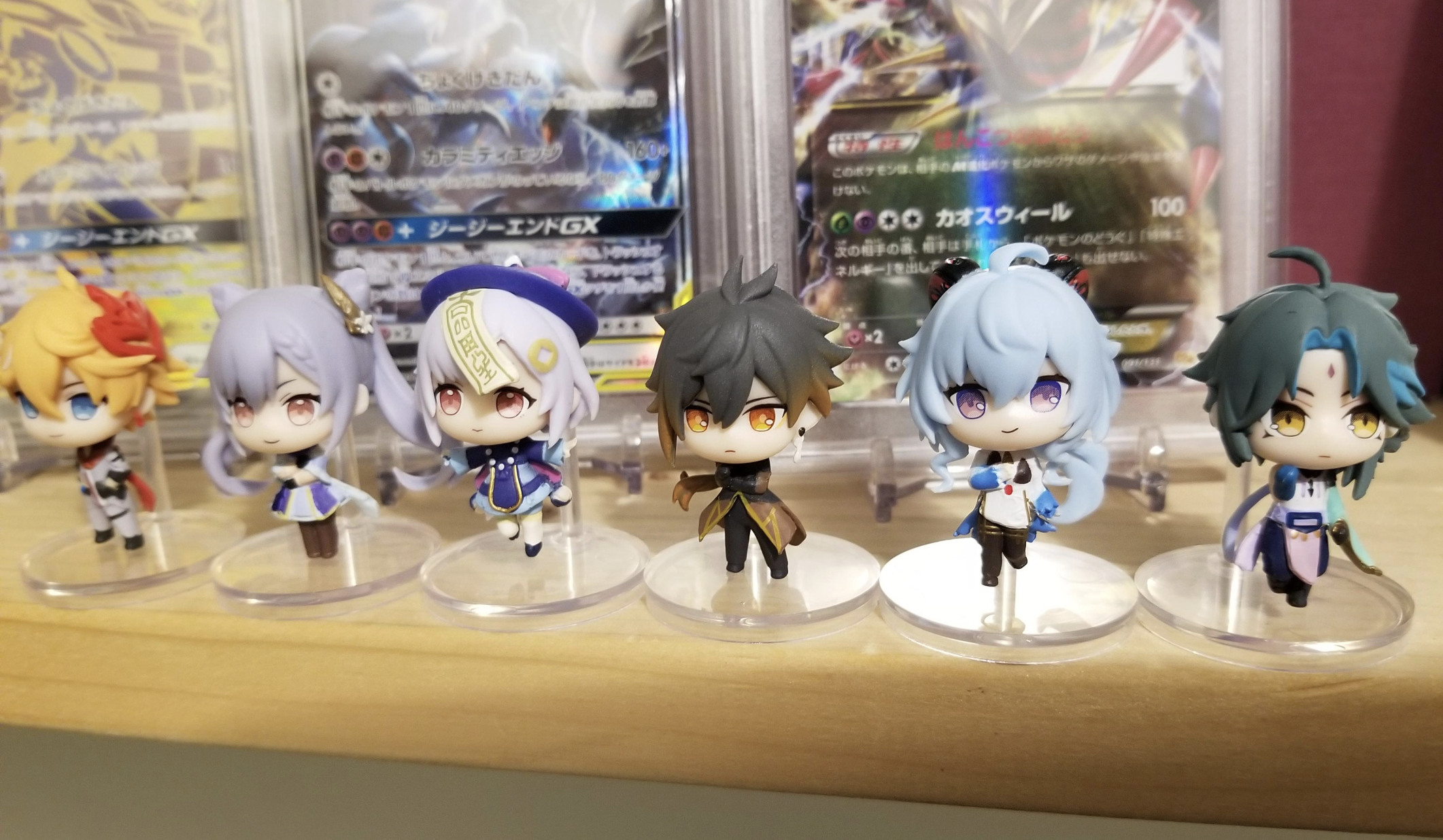 Six small anime-style character figurines are displayed on a shelf in front of trading cards, each with distinct colored hair and outfits, standing on clear round bases.