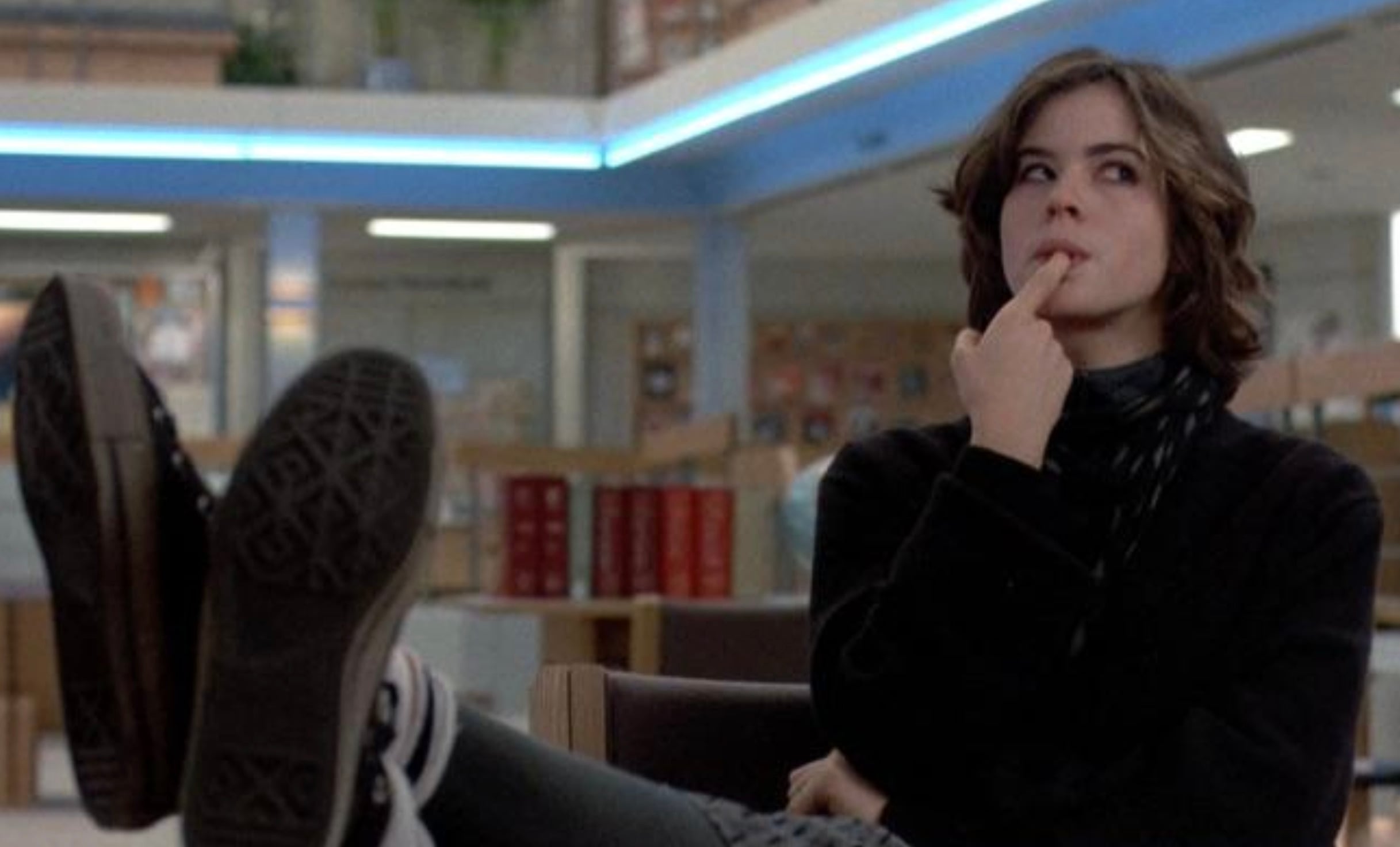 A person with short brown hair dressed in black sits with their feet up on a table, looking thoughtful and touching their lips with one finger in a spacious room with fluorescent lighting.