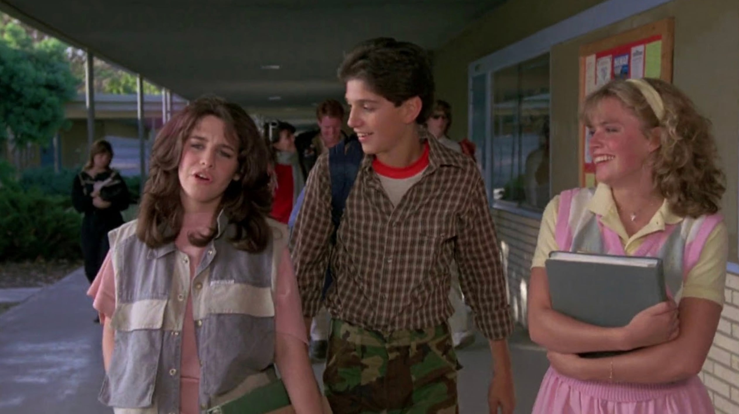 Three teenagers walk down a school hallway. The girl on the left looks frustrated, the boy in the center smiles at her, and the girl on the right, holding books, laughs. Other students and a bulletin board are visible in the background.