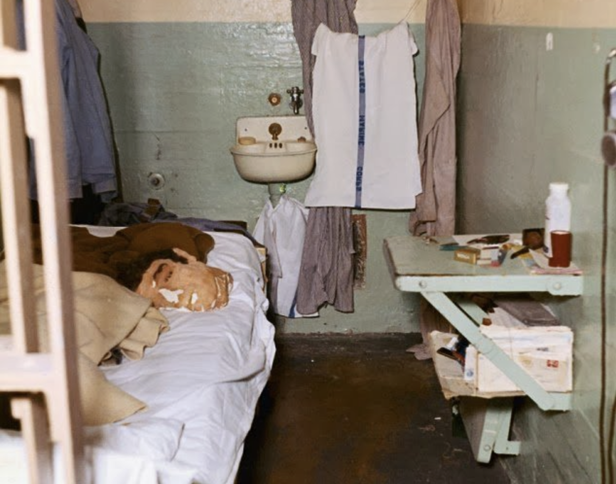 A small prison cell with a single bed, a cracked head mannequin on the pillow, a sink with clothes hanging above it, a green shelf with toiletries and books, and worn walls painted green and beige.