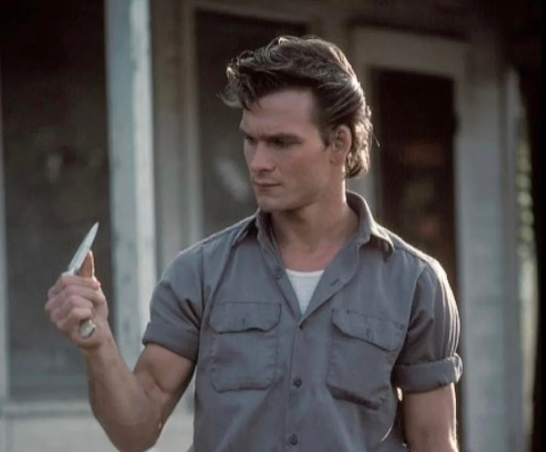 A man in a gray short-sleeve button-up shirt holds a knife in his right hand and looks at it intensely; he stands outdoors in front of a building with large windows.