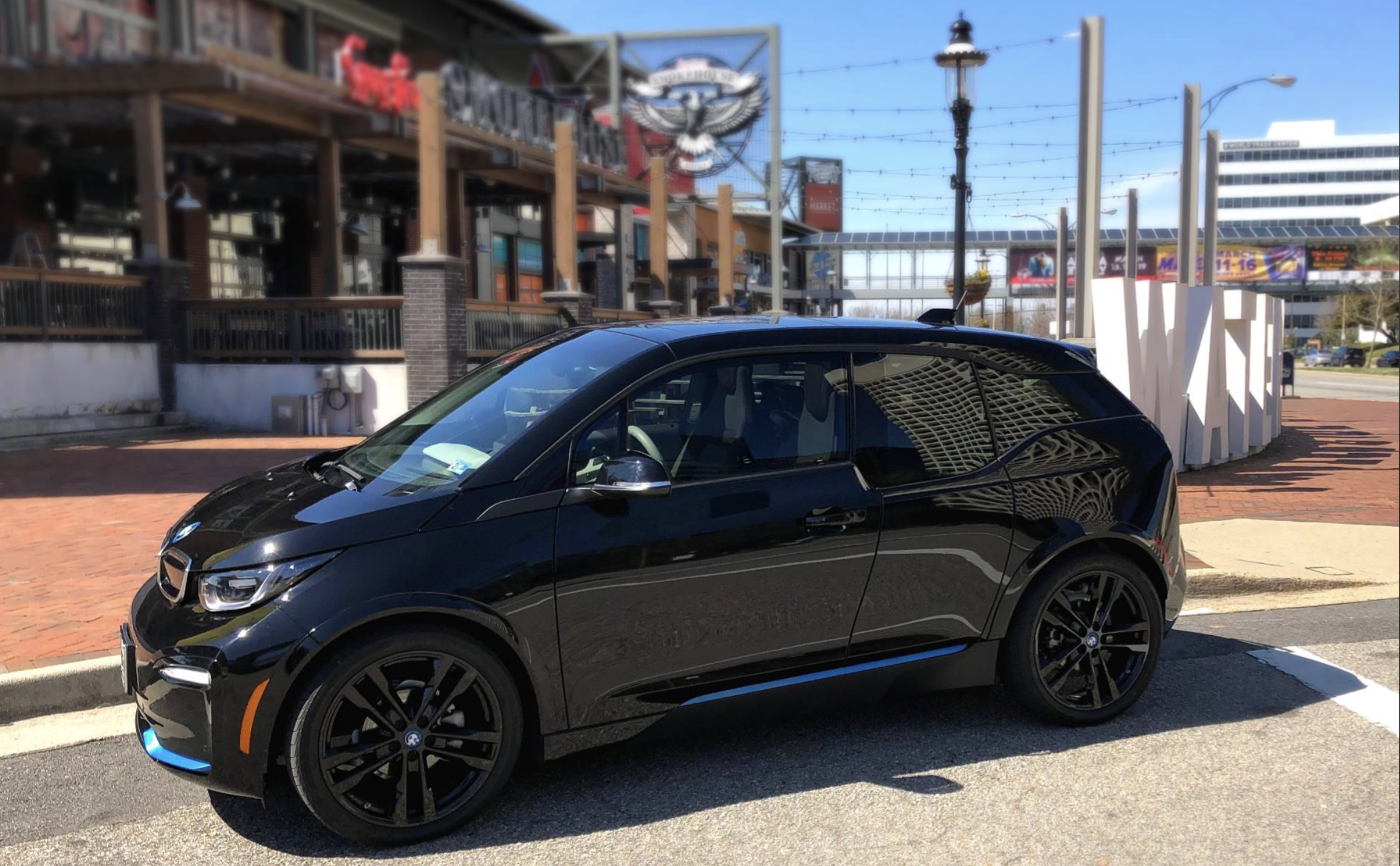 A black BMW i3 electric car is parked on a city street, with a modern building and outdoor restaurant visible in the background on a sunny day.