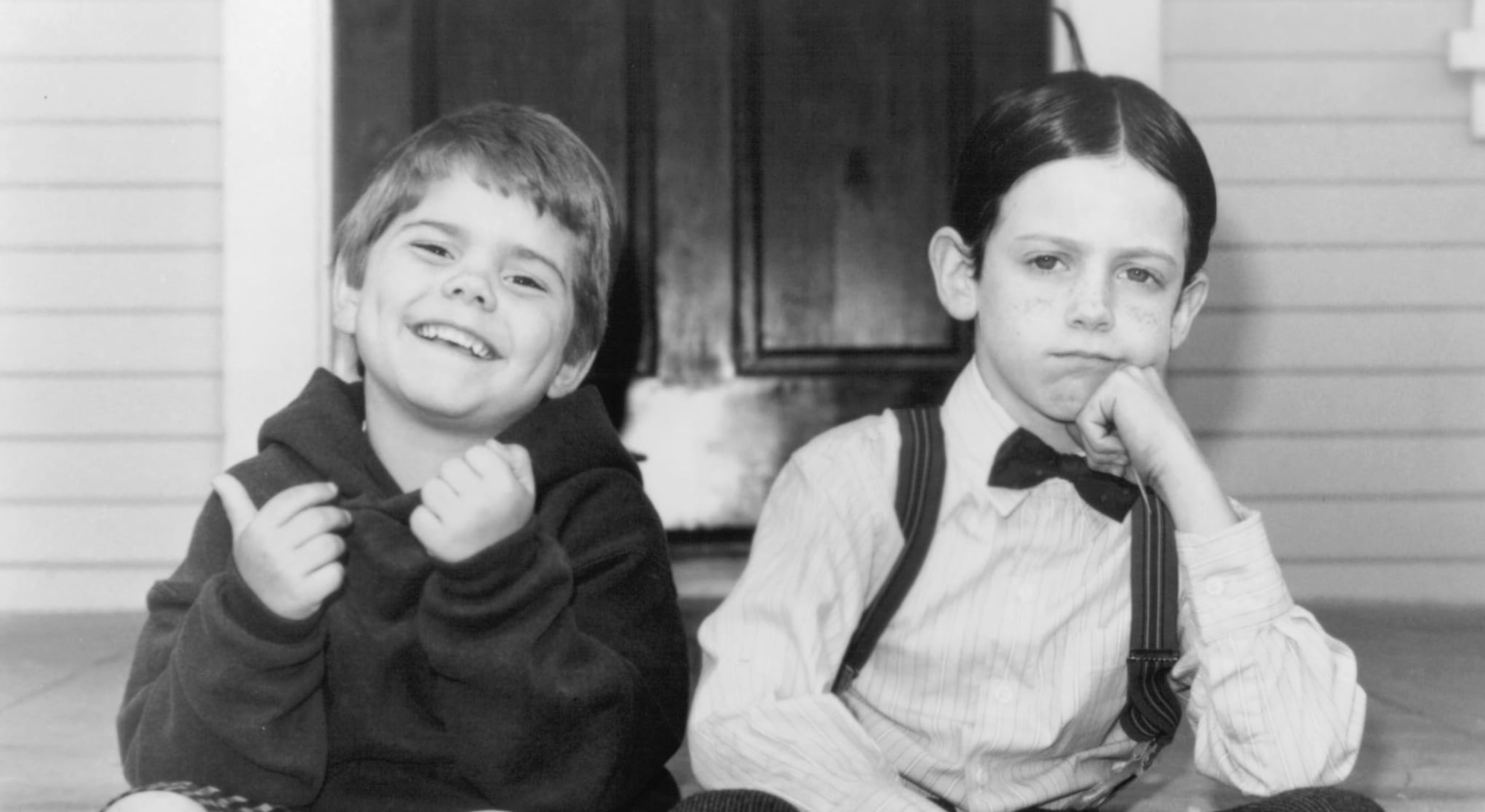 Two children sit on a porch; one is smiling and wearing a hoodie, while the other, dressed in a collared shirt, bow tie, and suspenders, looks serious with their chin resting on their hand.