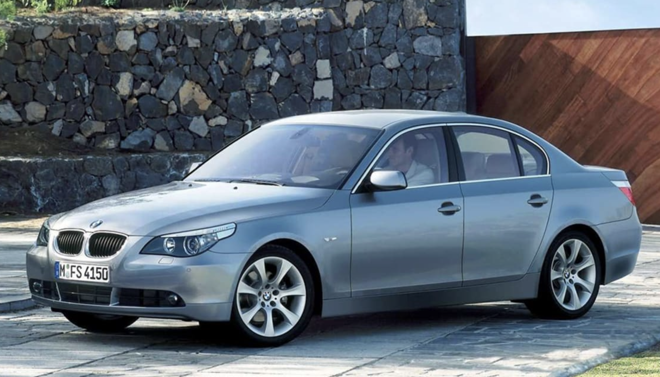 A silver BMW sedan is parked on a paved driveway with a stone wall and wooden paneling in the background. A person is seated in the driver’s seat, and the car is viewed from the front left side.