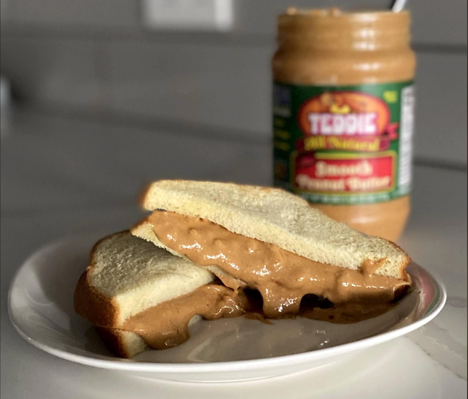 A peanut butter sandwich with thick, gooey filling sits on a white plate, with a jar of Teddie Smooth Peanut Butter in the background on a kitchen counter.