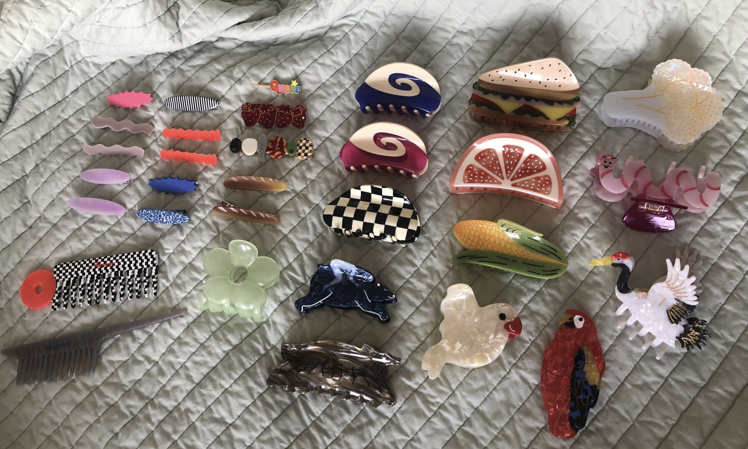 A collection of colorful, uniquely shaped hair clips, including clips shaped like food, animals, flowers, and abstract designs, arranged neatly on a textured, light-colored quilt.