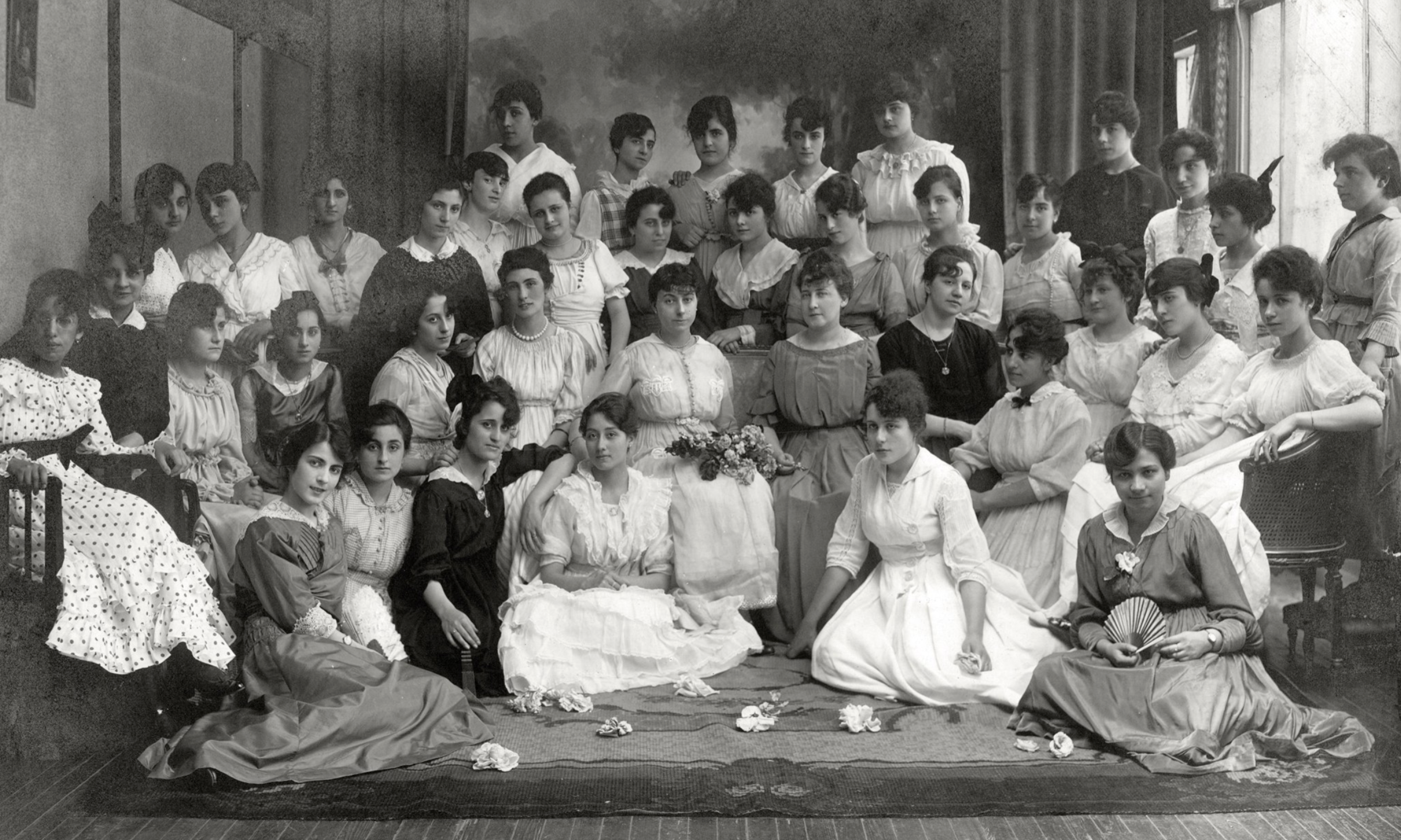 A black-and-white vintage photograph of a large group of women in formal dresses, posed indoors. Some are seated on chairs or on the floor, while others stand behind them, all facing the camera.