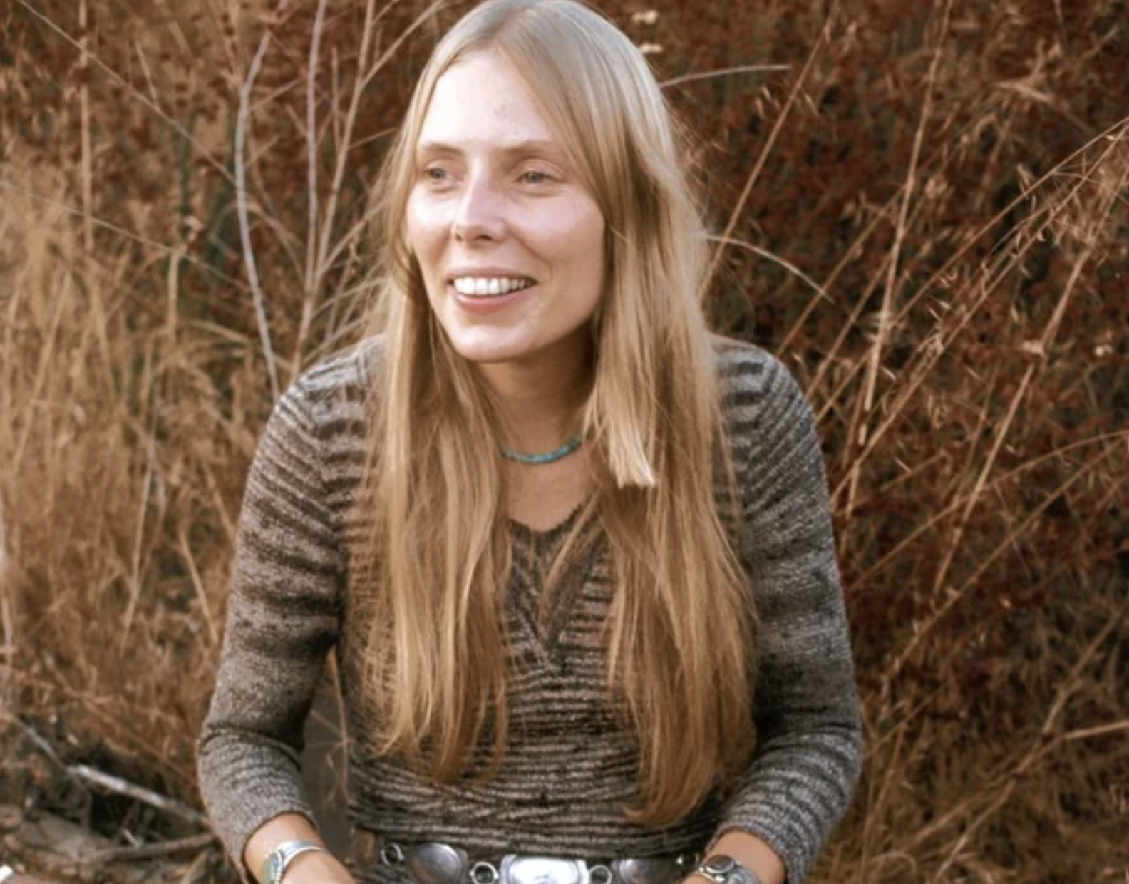 A woman with long blonde hair sits outdoors, smiling. She wears a striped long-sleeve top, turquoise necklace, large belt, and bracelets. Dry grass and plants surround her in a natural, earthy setting.