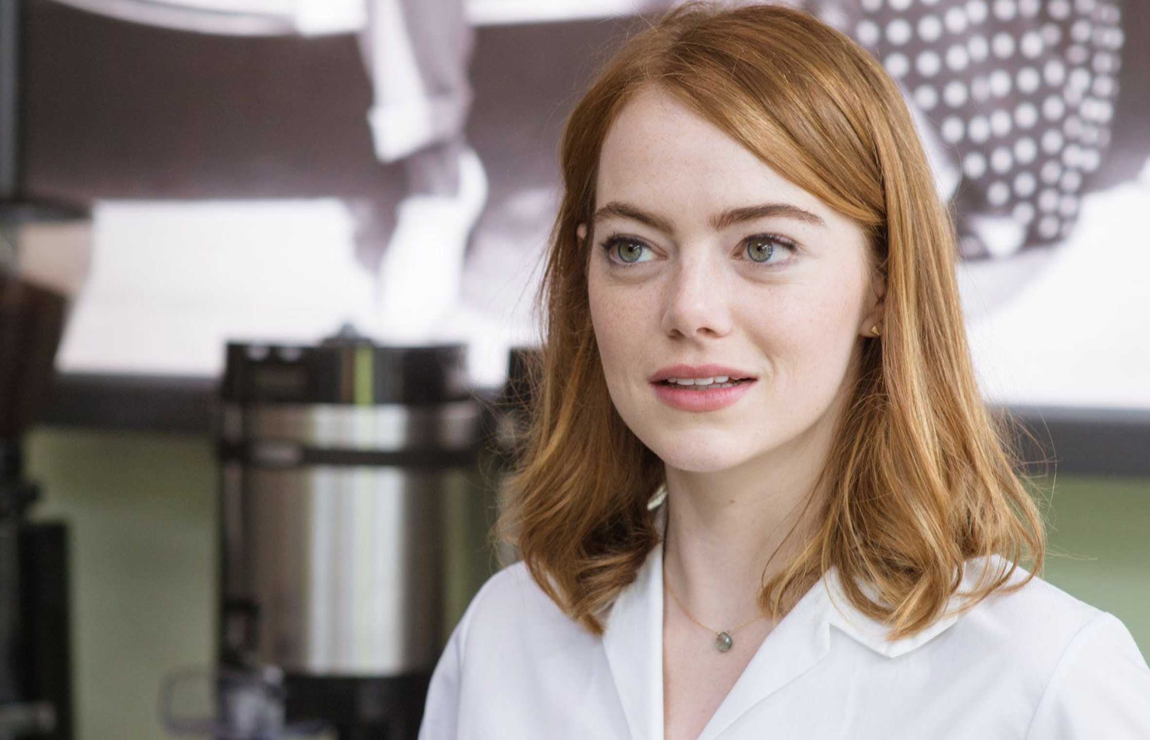 A woman with shoulder-length red hair and fair skin, wearing a white collared shirt and a delicate necklace, stands indoors with a coffee machine and a blurred black-and-white photo in the background.