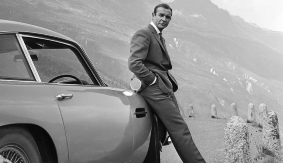 A man in a suit leans casually against a classic car parked on a mountain road, with a scenic view of mountains and valleys in the background. The image is in black and white.