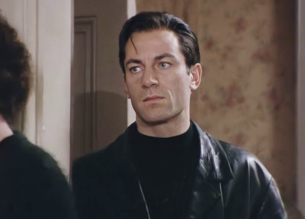 A man with slicked dark hair and blue eyes, wearing a black turtleneck and black leather jacket, stands indoors near a floral wallpapered wall, looking serious.
