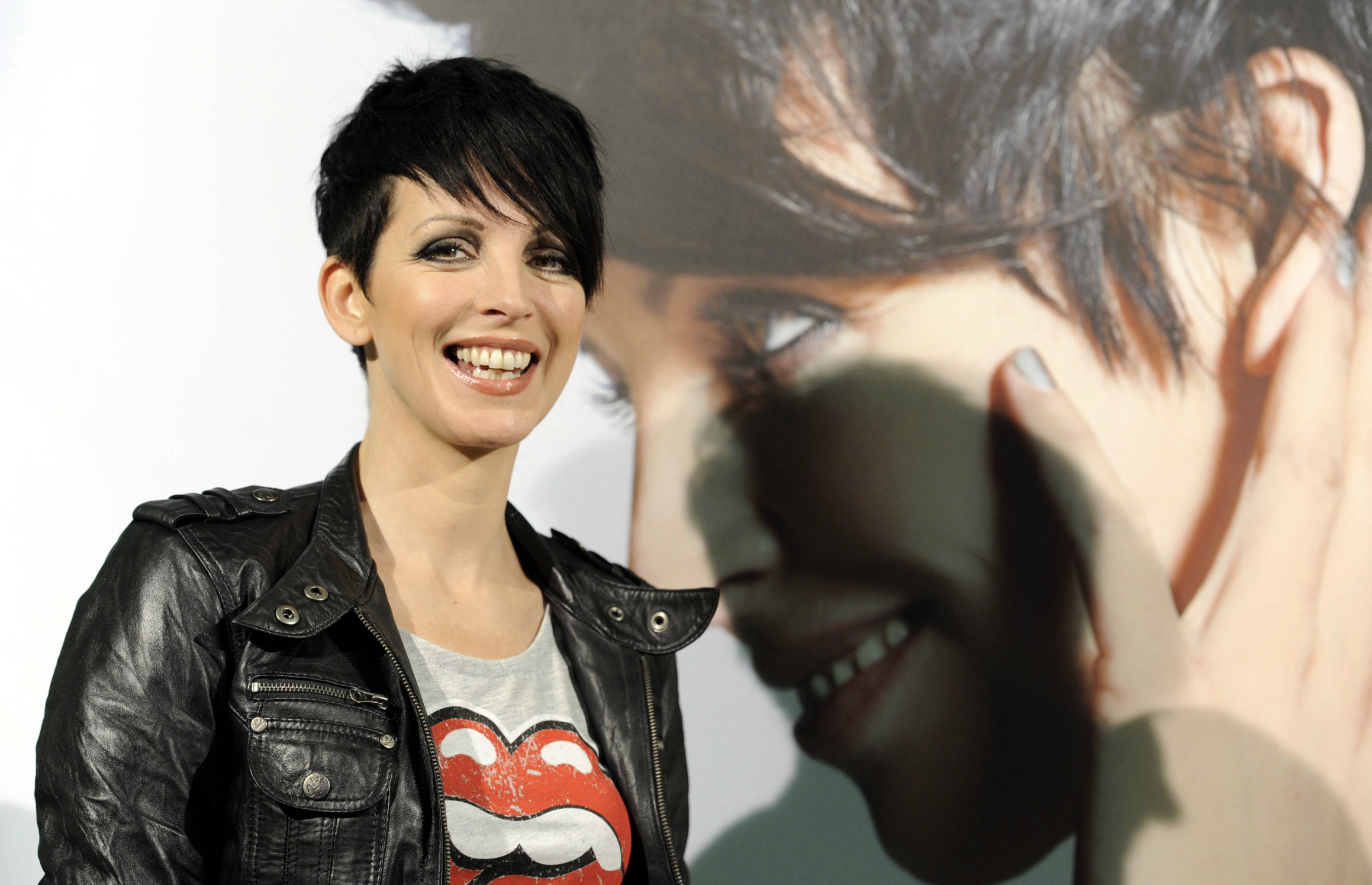 A woman with short black hair and a leather jacket smiles in front of a large poster featuring a close-up of her own face. She wears a T-shirt with a red lips graphic.