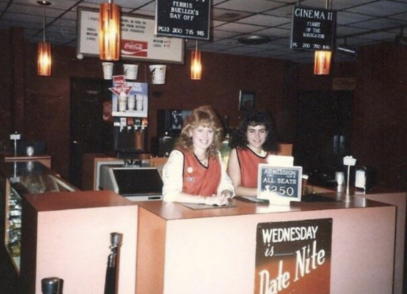 Two employees in red vests stand behind a concession counter at a retro movie theater with warm lighting. Signs advertise ticket prices and movies, and a &ldquo;Wednesday is Date Nite&rdquo; sign is displayed on the counter.